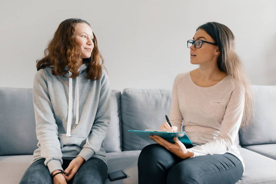 A teen in a grey hoodie sits on a couch while talking with a woman holding a clipboard in a pink shirt next to her